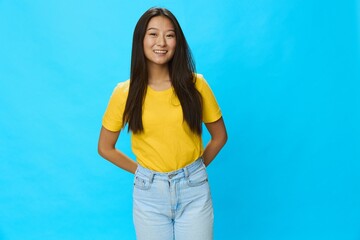 Asian woman smiling with teeth pointing finger at loose background in yellow T-shirt and blue jeans on blue background, copy space