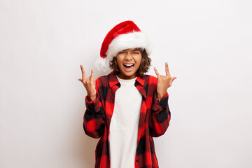 Fototapeta na wymiar Young dark skinned teenager, makes rock n roll sign, dressed in Santa Claus hat and red shirt, isolated over white wall