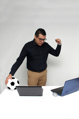 Latino adult office man watches football games on his work laptops during office hours in the morning, he sees him nervous, sad, stressed, angry next to his soccer ball
