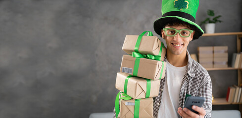 positive guy use his smartphone type text messages search St.Patricks day discounts wear leprechaun hat in house, holding many gift boxes