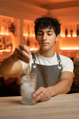Young male bartender in apron stirs alcohol cocktail with a special bar spoon.
