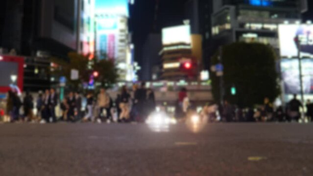 Many people walk and some run at pedestrian crossing, blurred version of footage. Lively Shibuya scramble crosswalk at evening or night. Bright headlights of cars flash through moving crowd