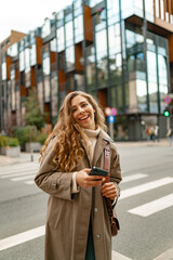 Fototapeta premium Smiling curly woman wearing warm coat walking down the street and using her phone