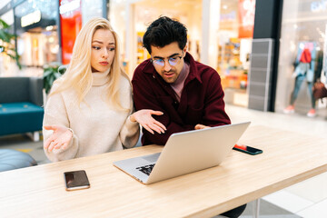 Puzzled young couple using typing laptop computer together, discussing purchase on the Internet, looking to screen sitting at table in hall of shopping mall with bright modern interior.