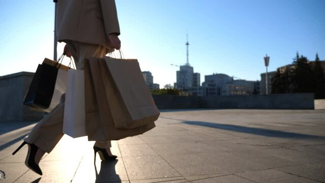 Young woman holds shopping bags walking at sidewalk after purchases. Elegant girl in suit carries paper packets going along sunny urban street. Concept of sales and discounts. Close up Slow motion