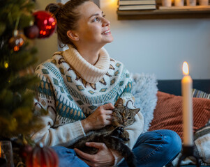 Young curly smiling woman in warm sweater with a cat with a Christmas toy while sitting at home on the couch next to the Christmas tree