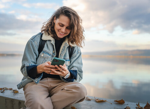 Pretty young woman in a jacket with a backpack using a smartphone while sitting on the promenade near the lake