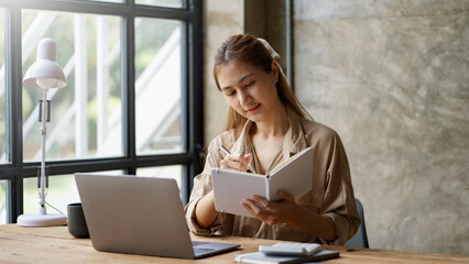 Young Asian businesswoman sitting happily working on her laptop and taking notes fluently and smiling happily at her assignment.