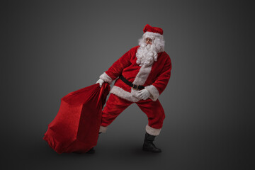 Studio shot of funny santa claus dragging big bag with gifts against gray background.