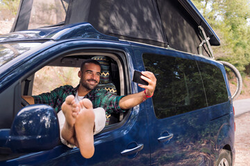 smiling young man taking a selfie with his phone sitting on the seat of his camper van with the feet leaning in the window, concept of nature travel and nomadic lifestyle