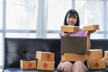 
Asian woman sitting on sofa and working at home, she is excited and smiling holding packaging box to write customer address in mailbox to send online sale items. The concept of SME business owners