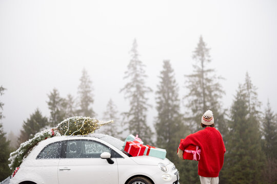 Young woman in red sweater enjoys great view on pine forest, while traveling by car in mountains during winter holidays. Vehicle with Christmas tree, wreath and gift boxes. Image with copy space