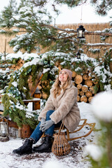 A cute girl in a light fur coat sits on a sled under a pine tree in the snow
