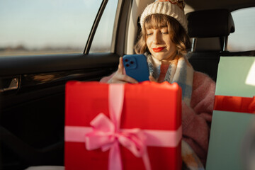 Young woman dressed for the holidays drives in car on back seat with beautifully wrapped Christmas presents and using phone. In anticipation of the winter holidays, preparation and shopping concept