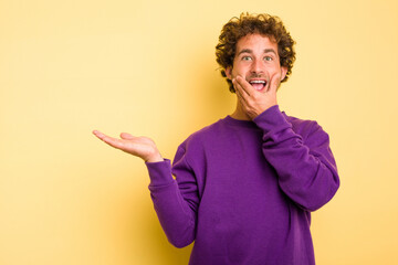 Young smart caucasian man on yellow background holds copy space on a palm, keep hand over cheek. Amazed and delighted.