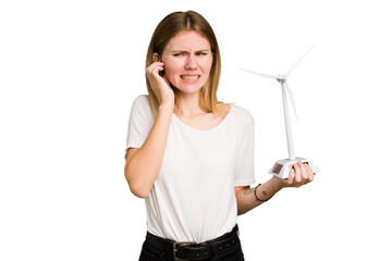 Young caucasian woman holding a small wind energy mill isolated covering ears with hands.