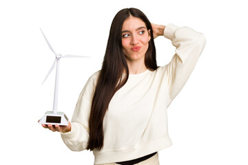 Fototapeta premium Young caucasian woman holding a small wind energy mill isolated touching back of head, thinking and making a choice.