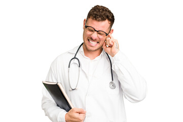 Young doctor caucasian man holding a book isolated covering ears with hands.