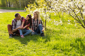 Naklejka na ściany i meble Happy parents mom and dad, daughter, young family outdoors in spring against the background of blooming apple and cherry trees
