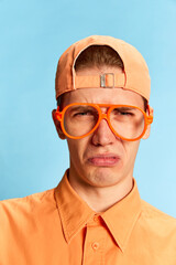 Closeup portrait of emotional young man in eyewear and cap looking at camera with vivid emotions isolated over light blue background. Mental health, facial expression, emotions concept