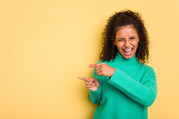 Young Brazilian curly hair cute woman isolated on yellow background pointing with forefingers to a copy space, expressing excitement and desire.