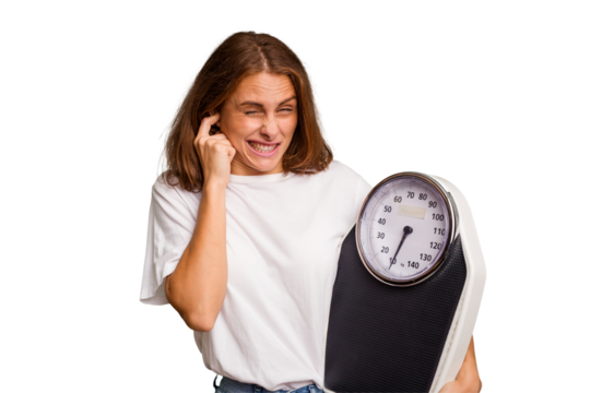 Young caucasian woman holding a scale and measure tape isolated covering ears with hands.
