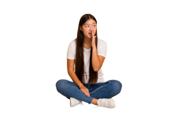 Young asian woman sitting on the floor cutout isolated is saying a secret hot braking news and looking aside