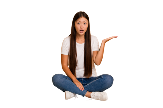 Young asian woman sitting on the floor cutout isolated impressed holding copy space on palm.