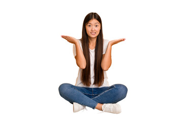 Young asian woman sitting on the floor cutout isolated confused and doubtful shrugging shoulders to...