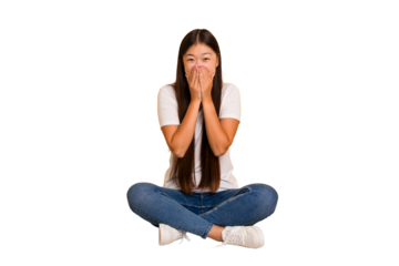 Young asian woman sitting on the floor cutout isolated laughing about something, covering mouth with hands.