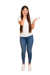 Young asian woman standing, full body cutout isolated holds copy space on a palm, keep hand over cheek. Amazed and delighted.