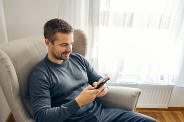 A man is relaxing in his chair, typing messages on social medial while smiling at the phone.