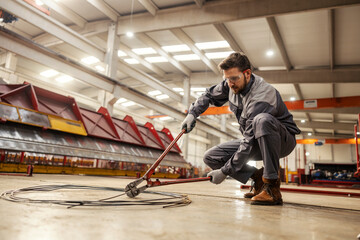 A heavy industry worker is cutting wire with big wire cutter while crouching in facility.