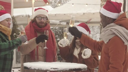Multiethnic friends in red Santa hats toasting with champagne glasses while celebrating Christmas holidays in cozy village cafe decorated with lights and wreaths