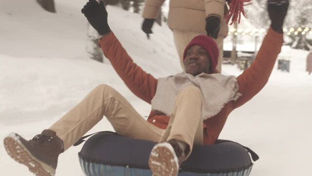Young cheerful Black man having fun while riding snow tube carried by his friend along snowy path in winter forest