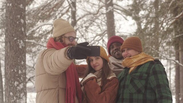 Group of young diverse friends taking selfie portrait on smartphone outdoors in winter forest