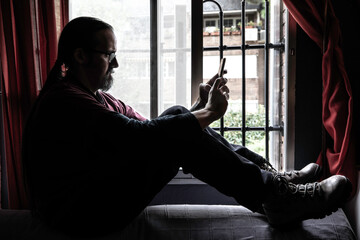 Adult man with ponytail and beard checks his mobile phone sitting on the back of a sofa against a window. Moody style