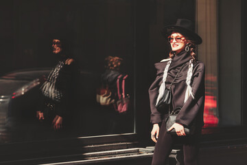Stylish young woman model in sunglasses and white braids walking on city street wearing black...