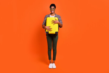 Full length body size view of attractive cheerful trendy girl holding hugging suitcase isolated over vivid orange color background