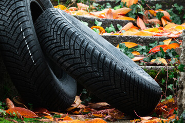 Fototapeta na wymiar Pair of brand new all weather tires lying on wet asphalt covered in fallen golden leaves in rainy autumn season.