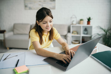 smiling brunette girl typing on laptop near notebook on table at home