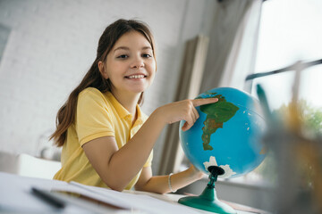 cheerful girl pointing at globe and smiling at camera during homeschooling on blurred foreground