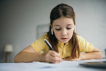 Naklejka na ściany i meble preteen girl in wireless earphones writing in notebook while doing homework