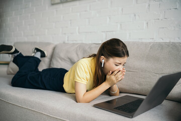 bored girl in wireless earphone yawning near laptop on comfortable couch in living room