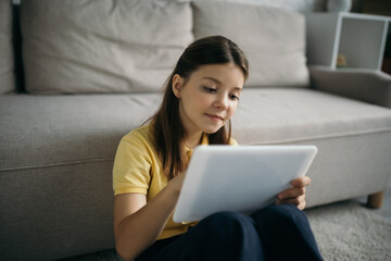 positive preteen girl using digital tablet near blurred couch in living room