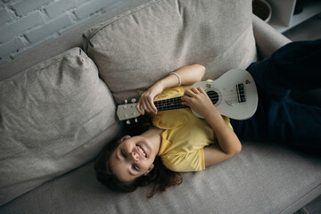 top view of joyful girl lying on sofa and playing small hawaiian guitar
