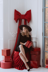 Beautiful elegant woman in a black holiday dress and hat among a lot of red New Year's gifts. Soft selective focus.