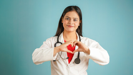 Portrait of a young female doctor cardiologist showing heart symbol with fingers, Cheerful Asian Indian woman doctor in apron and stethoscope isolated blue studio background, expressing love and care