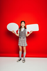 full length of girl in checkered skirt and gumshoes holding blank speech bubbles on red background