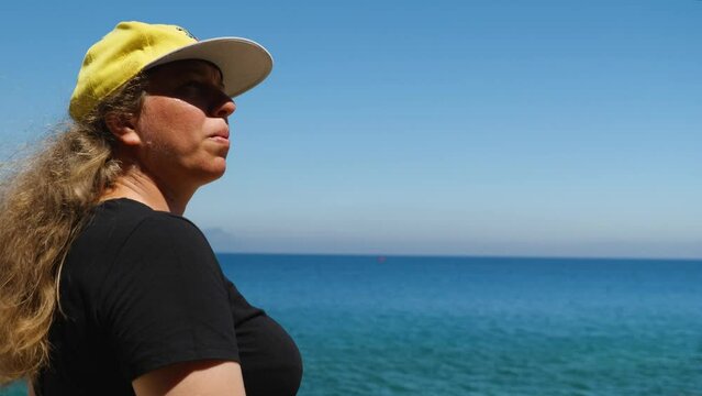 Girl in black jersey and baseball cap walking along the promenade along the sea. The camera is moving in parallel. There is a beautiful sea and mountains in the background. Close-up